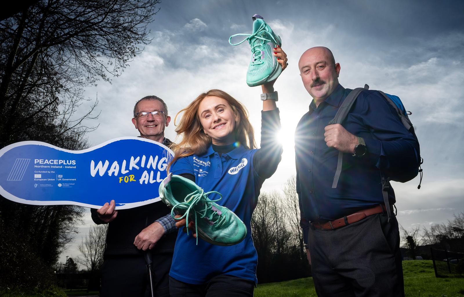 Three people standing outdoors in a park. The person in the centre, wearing a blue ‘Walk for All’ shirt, holds up a pair of turquoise walking shoes. One person on the left holds a blue sign reading ‘PeacePlus Northern Ireland – Walking for All,’ and the person on the right wears a backpack. Trees and a cloudy sky are visible in the background, with sunlight behind the group.